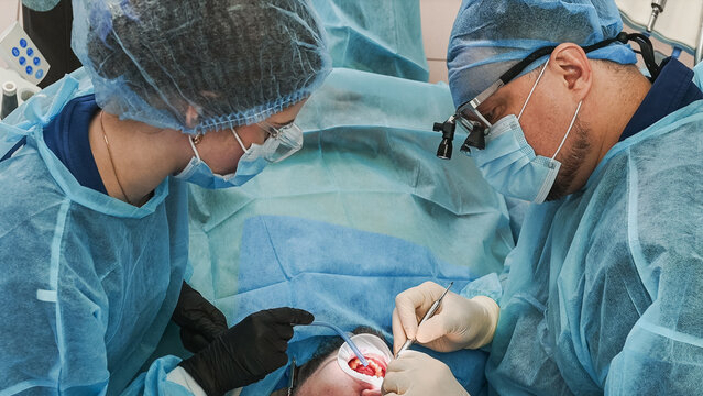Dental specialists restoring gum tissue and placing dental implants, working inside patient mouth with sterile instruments during oral surgical procedure