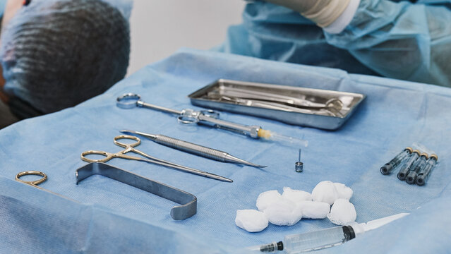 blurred close-up of surgical instruments laid out on a sterile drape on a table. A surgeon, faceless and unrecognizable, is in the background. An orthopedic dentist performs a dental implant.