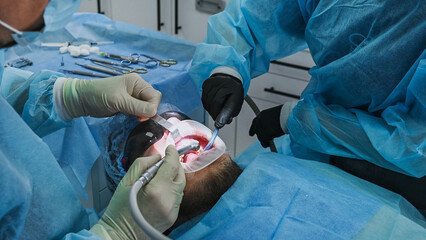 Dental surgeon and female dental assistant perform dental implant procedure, with assistant using saliva ejector while dentist works on patient in chair wearing sterile gown, gloves and cap. © illustrissima