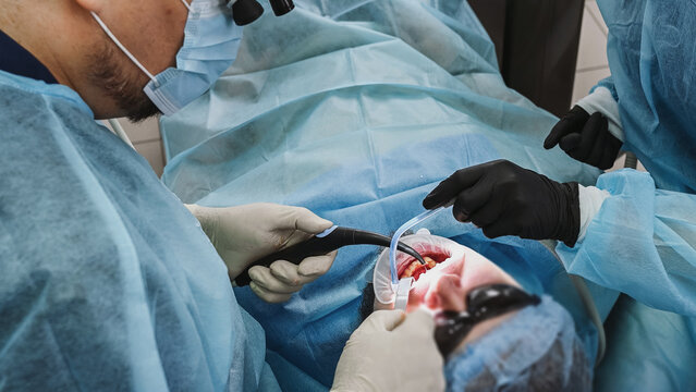Dental surgeon and female dental assistant perform dental implant procedure, with assistant using saliva ejector while dentist works on patient in chair wearing sterile gown, gloves and cap.