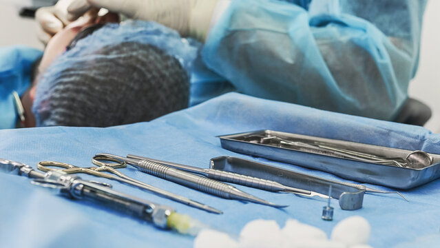 Sterile dental instruments displayed on surgical table in foreground, while faceless dentist performs implant placement on patient in blurred background during oral surgery