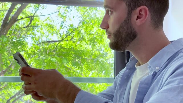 Young handsome man with a beard using a mobile phone, texting, scrolling, and browsing online while sitting near a window with a green, natural background of trees and foliage