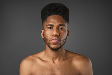 Obraz premium Beauty portrait of concentrated young African man looking at camera, shirtless and isolated over gray background, confident male model, skincare, body care, wellness and masculine lifestyle concept