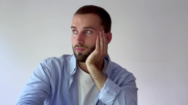 Handsome caucasian man with a beard looking bored and uninterested, propping his head on his hand while sitting in a studio with a plain white background, expressing apathy and laziness