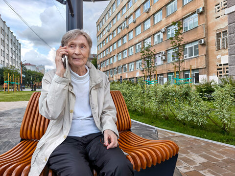 Elderly woman talking on smartphone while sitting on bench in city park. Senior citizen receiving phone call with serious expression, phone scam fraud target senior citizen