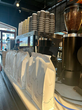 A coffee shop counter features numerous white paper bags and stacked coffee cups. A coffee machine and grinder are prominently displayed, along with warm light creating a cozy atmosphere.