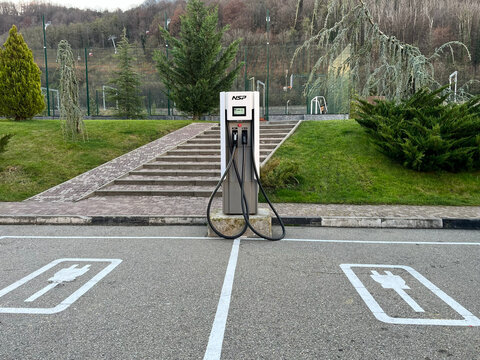 An electric vehicle charging station stands in a parking lot surrounded by greenery. Charging cables hang from the unit. The area is well-maintained and spacious.