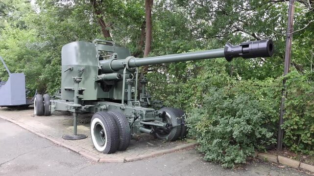 View of a historic anti-aircraft gun displayed in a park setting with lush green trees and a paved path in Vladivostok, Primorsky Krai, Russia.