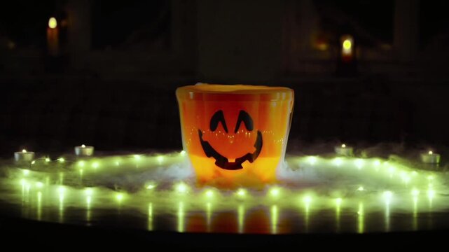 
Sebuah ember plastik berwarna oranye terang berbentuk labu&mdash;yang menyerupai *jack-o'-lantern* Halloween dengan wajah menyeringai jahat&mdash;terletak di tengah lingkaran lampu hias (*fairy lights*).