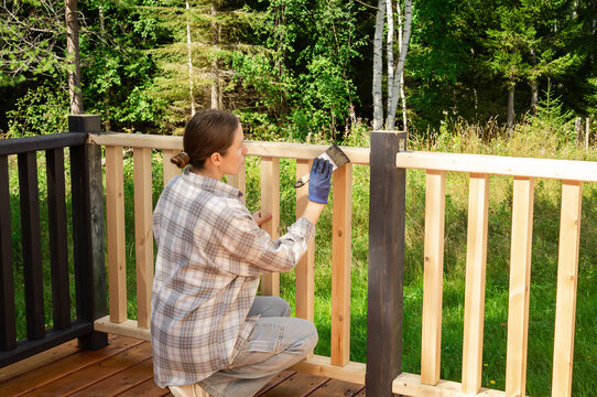 Woman in a checkered shirt applying protective wood stain or oil to a terrace railing. Handy woman using paintbrush for DIY home improvement, backyard deck maintenance and renovation project.