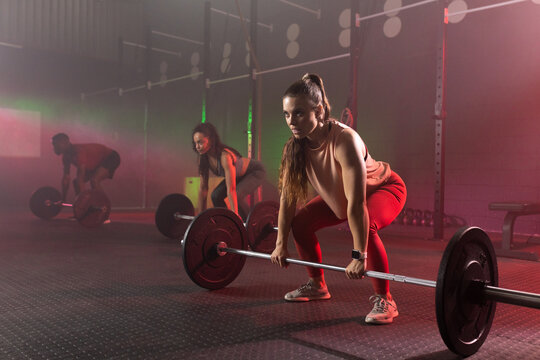 Diverse classmates wearing athletic clothing lifting loaded barbells in dim gym with kettlebells