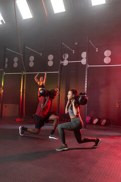 Diverse workout group performing black sandbag lunges and hanging pulls at functional gym with rig