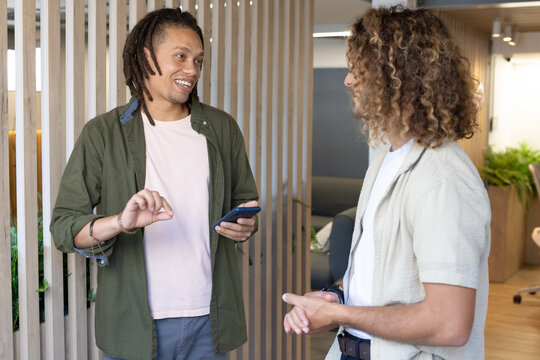 African American male coworkers talking in office lounge by wooden slats holding smartphone