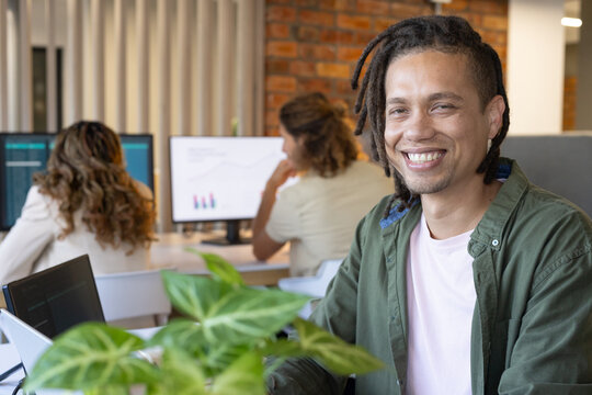 Diverse coworkers working in open-plan office, using monitors, laptop, keyboard, potted plant