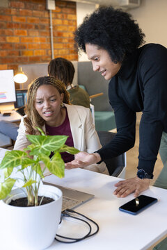 African American coworkers discussing project at office desk with silver laptop, plant, copy space