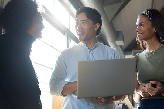 Diverse coworkers collaborating around laptop at office by grid window with smartphone
