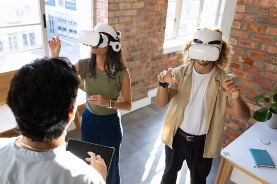 Colleagues wearing white VR headsets and reaching forward in bright office near tablet and notebook