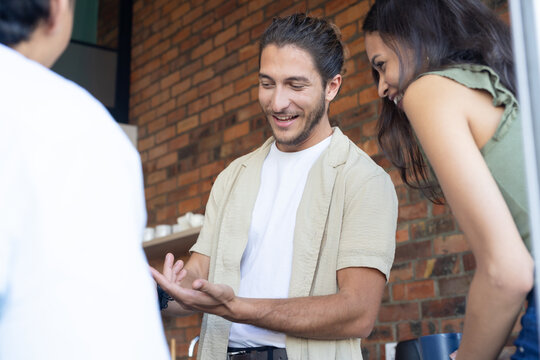Diverse coworkers standing at cafe counter, man in beige overshirt gesturing to open palm near mugs