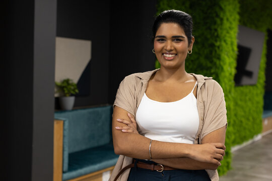 Indian woman standing with arms crossed, smiling in office lounge with living wall, beige shirt