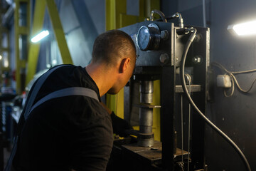 Drill press stands near yellow frame inside workshop corner. Male technician adjusts spindle drilling metal part while checking alignment by eye © SlavaStock