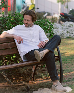 A young man is sitting on a wooden park bench with his legs crossed. He is wearing a short-sleeved white shirt and dark trousers.