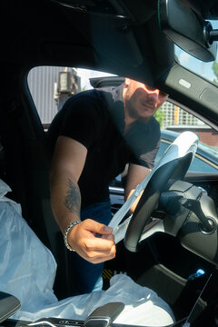 Open car door frames steering wheel and dashboard in sunlight. Male technician stretches protective cover tucking edge under rim for secure fit