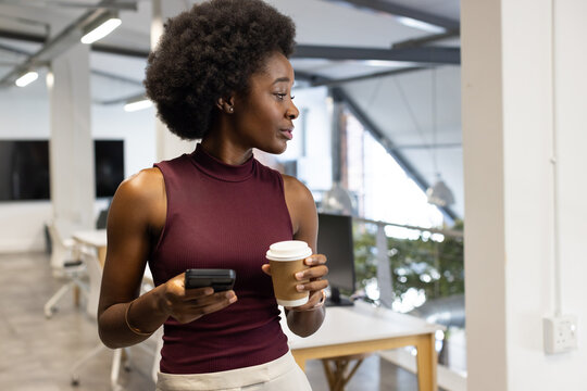 African adult woman standing holding phone and to-go cup in office in maroon top looking right