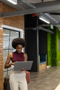 African woman standing in modern office wearing burgundy top and beige trousers holding laptop