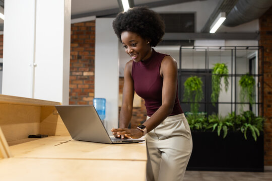 Adult African woman leaning on counter at office, using laptop and smartphone, wearing maroon top