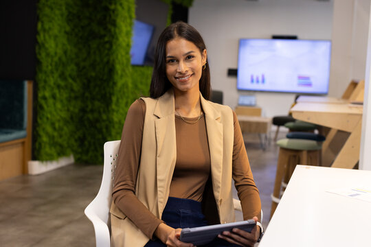 Female worker wearing blazer sitting on chair in lounge holding tablet viewing screen charts