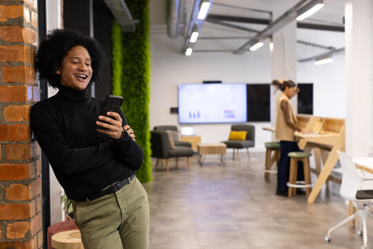 African American colleagues leaning on brick column, smiling with phone in office, copy space