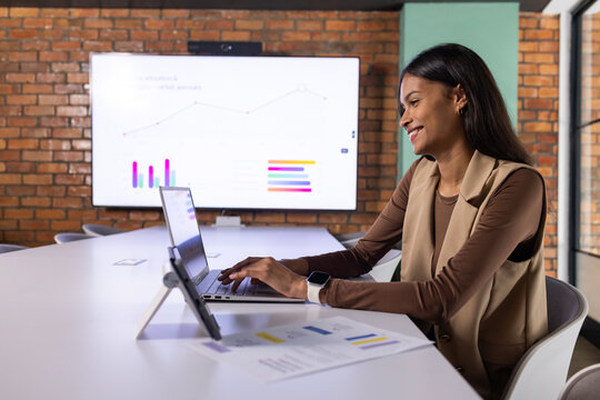 African American woman in blazer working on laptop with tablet-stand at table and charts