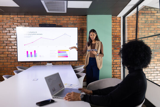 African-American woman in tan blazer with tablet presenting data on meeting-room screen, copy space