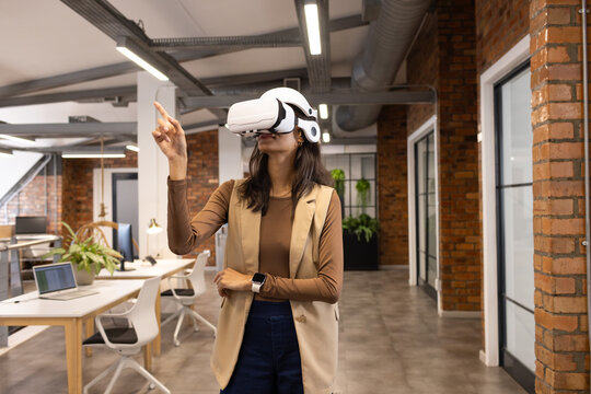 African American woman standing in modern open-plan office wearing white VR headset pointing upward