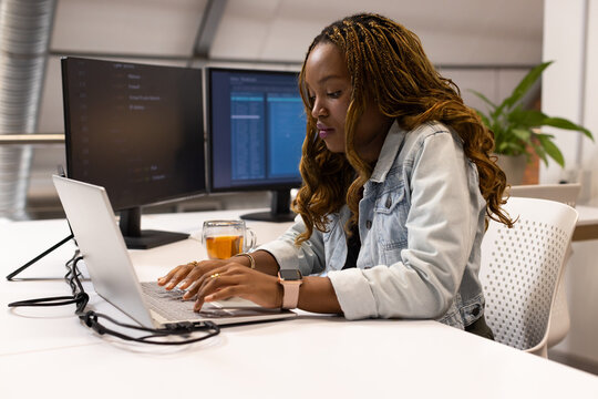 African American woman in denim jacket typing on laptop in open office with monitors, copy space