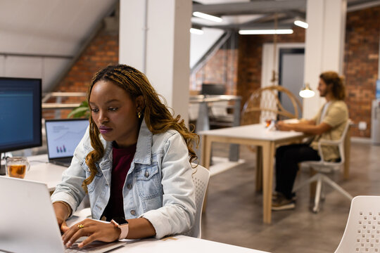 Diverse coworkers working in loft office with African American woman in denim using laptop, mug