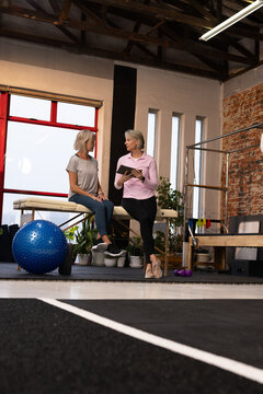 Female trainer and client in athletic clothes discussing plan at Pilates gym using tablet on plinth