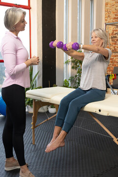 Mature coach guiding female client in gray tee doing seated raises with dumbbells on therapy table