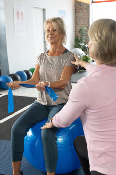 Senior females in sportswear, trainer guiding client on stability ball in studio stretching band