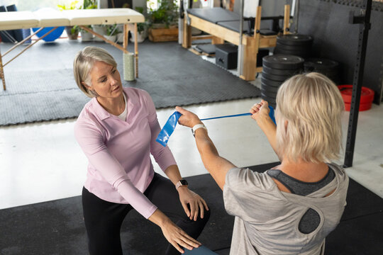Female trainer kneeling guiding client pulling blue resistance band on black mat in rehab studio