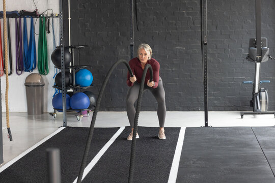 Woman in maroon top, gray leggings holding and oscillating heavy battle ropes in studio