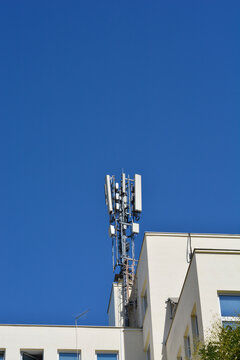 A low angle view shows a cellular network base station with multiple antennas on a modern building roof against a clear blue sky.