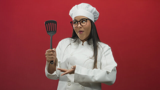 Hispanic chef woman holding spatula with right hand and presenting it with open left palm against a red studio backdrop; playful curiosity.