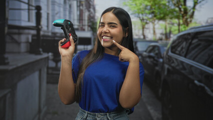 Woman holding a gamepad and points finger to cheek while smiling with controller raised in a street scene near parked cars and buildings  joyful play. © Krakenimages.com