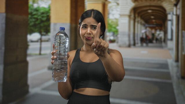 Woman holds water bottle and points index finger in arcade corridor, giving a direct hydration cue; hydration reminder warning.