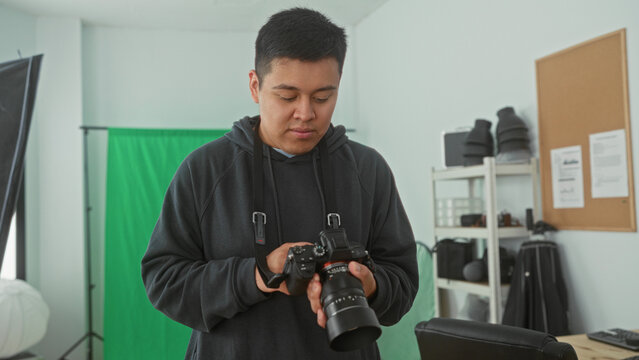 Man in hoodie adjusts camera controls and holds dslr in studio with green screen while checking settings; concentration craft.