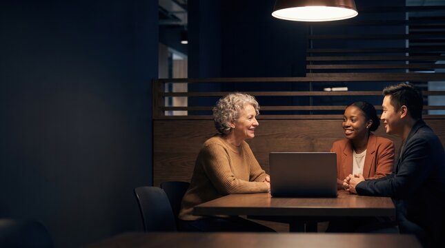 Multigenerational colleagues laughing during business meeting around laptop in modern office, sharing ideas and building teamwork in warm evening light