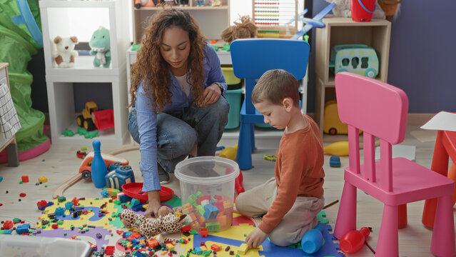 Woman kneeling, hand reaching for plastic blocks as young boy sorts toys on a playroom floor inside a building; learning calm.