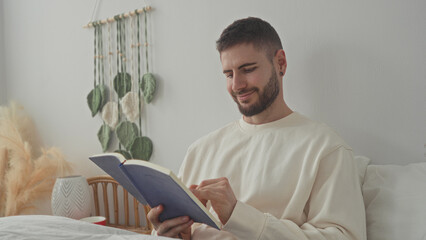 Obraz premium Man sits propped on bed reading and turning pages with bare hand while holding a blue paperback book near rattan headboard in building; calm focus.