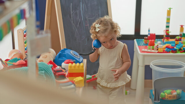 Little blonde girl holds blue toy telephone to her ear and dials beside a chalkboard and table of blocks in a playroom building; curiosity and play.
